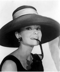 Un bel primo piano di Audrey Hepburn con un bel cappello e degli orecchini mentre tiene in bocca la stecca di un paio di occhiali. 