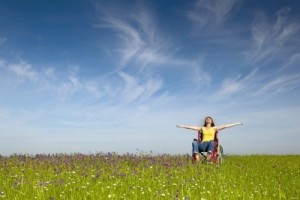 Una giovane donna in sedia a rotelle, con le braccia spalancate, su un prato fiorito. Alle sue spalle, un cielo quasi sereno.