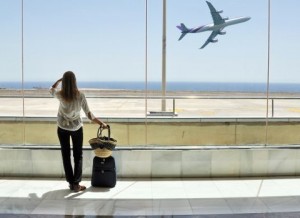 Una donna con bagaglio a mano attende di prendere l’aereo. La donna è fotografata in piedi, di spalle mentre osserva, attraverso una vetrata, un aereo appena decollato.